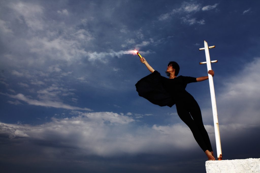 The WIM Round 2 Prompt photo features a woman dressed in black holding onto a white pole with her left hand while extending her body away. She holds a smoking flare in her right hand, lifting it high.