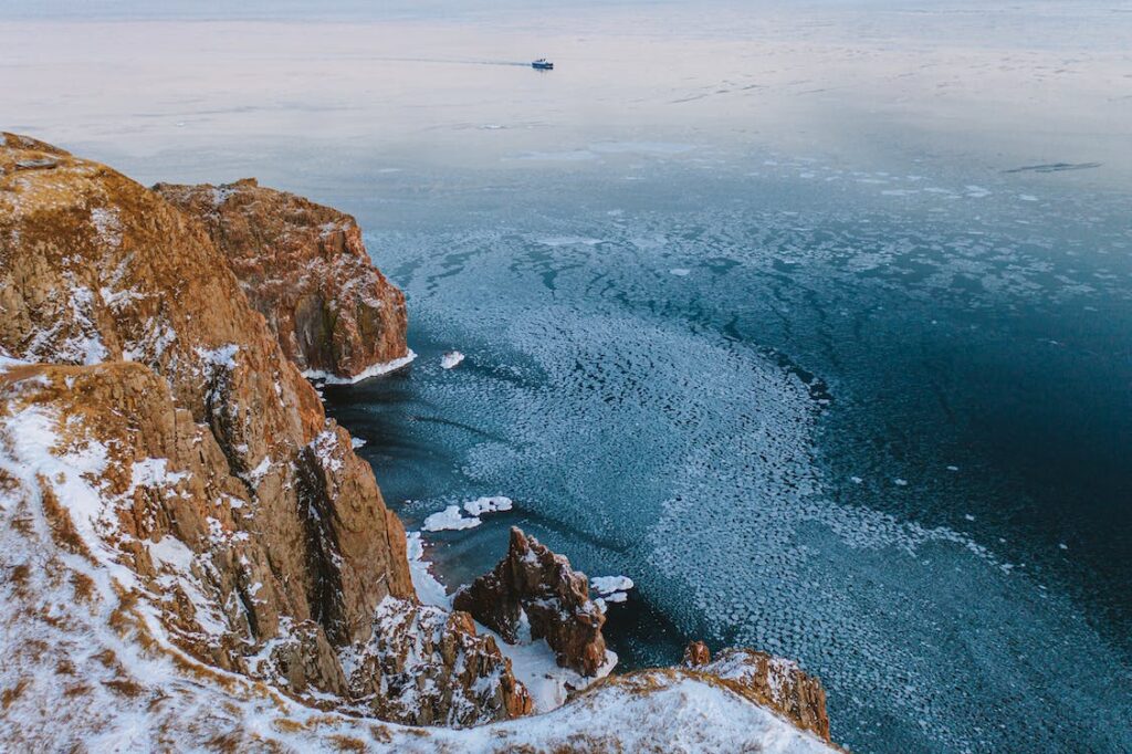 A snow-dusted brown cliff plunging into an ice-covered blue sea with a ship on the horizon